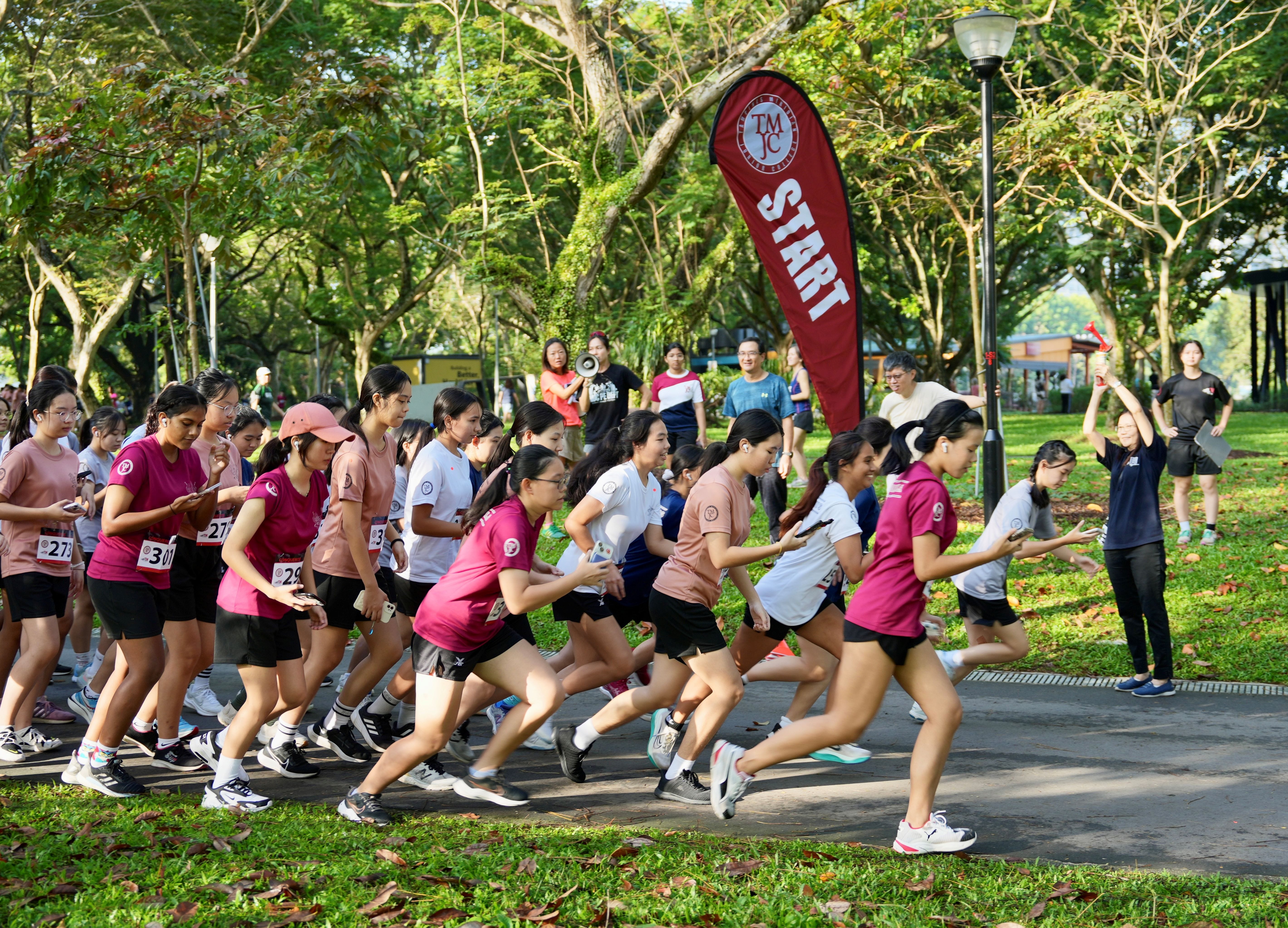 A large crowd of students in maroon and white shirts burst from a start line at an outdoor run event, with a red "START" banner visible among the trees.